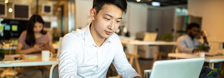 An Asian man working on a laptop in a cafe setting.