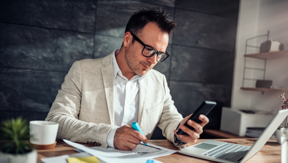 A Caucasian male wearing a beige suit in an office setting, sitting at a desk with a laptop opened in front of him. He is holding a mobile phone in his left hand and a blue highlighter in his right hand.