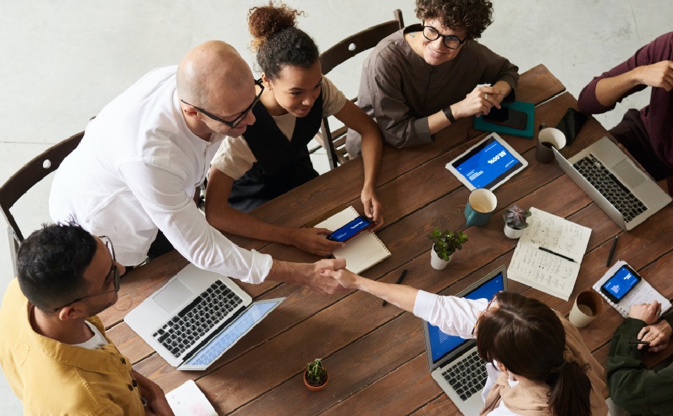 A top-down view of people sitting around a table with their laptops and mobile phones open. A male and a female are shaking hands across the table too.