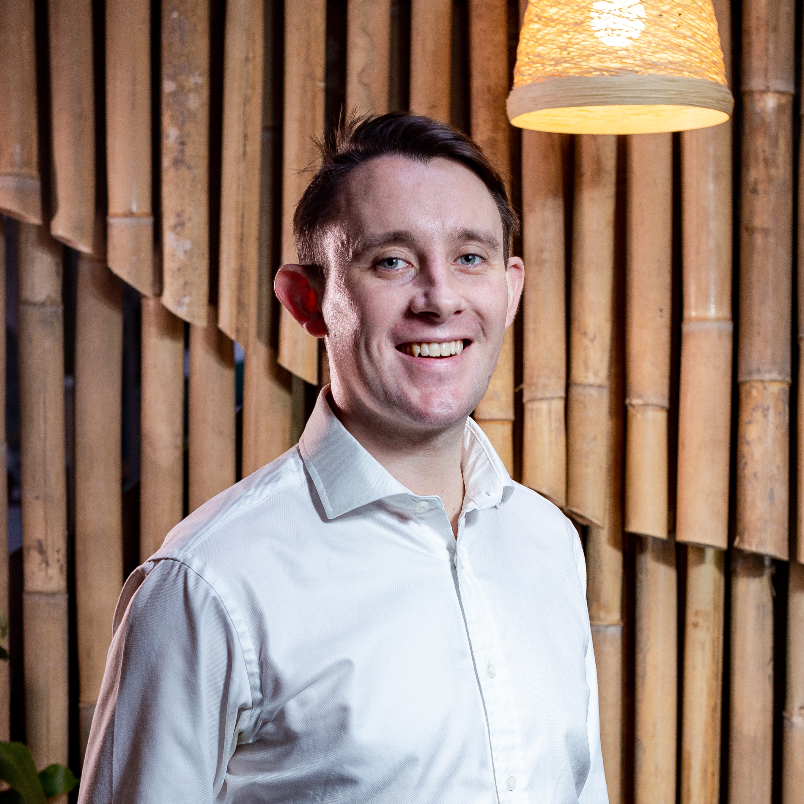 Smiling person in white shirt with bamboo background and hanging lamp.