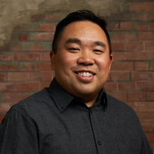 Smiling person in a dark shirt standing against a brick wall.