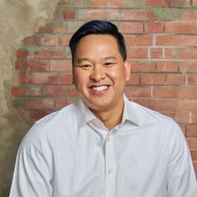 Man smiling in front of a brick wall, wearing a white shirt.