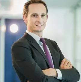 Man in a suit with a purple tie, arms crossed, smiling in a modern office.