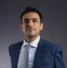 Man in a navy suit with a light tie stands against a gray background.