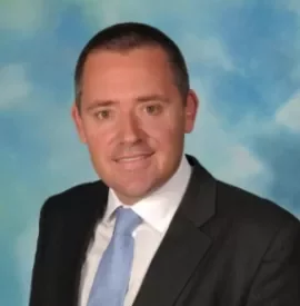 Man in a suit with a blue tie, smiling in front of a cloud-like background.