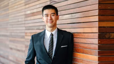 Man in a suit leaning against a wooden wall, looking at the camera.