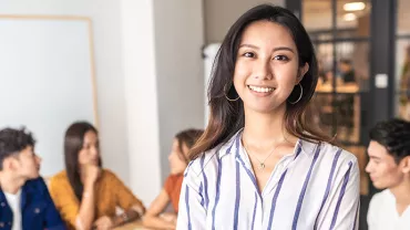 Smiling woman in a striped shirt stands in front of a group of people in a modern office environment.
