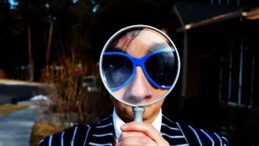 Person in blue sunglasses holding magnifying glass, outside on a sidewalk.