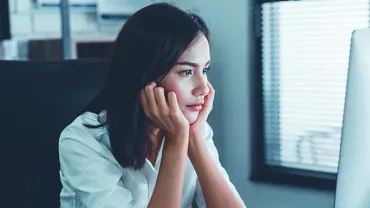 Woman in white shirt looking at computer screen with chin in hands.