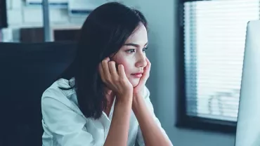 Woman in white shirt looking at computer screen with chin in hands.