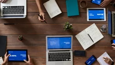 Top view of a group of people working with laptops, tablets, and notes around a wooden table.