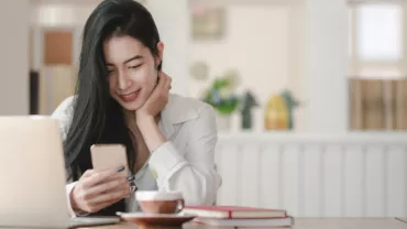 Woman smiling at phone in a cafe with a laptop and coffee.