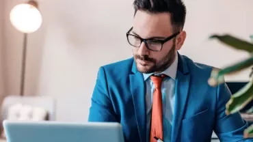 Man in a blue suit and red tie working on a laptop in an office.