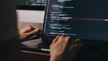 Close-up of hands typing code on a laptop in a dimly lit room.