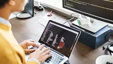 Person working on a laptop with code on the screen, seated at a desk with a large monitor.