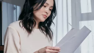 Woman with dark hair reads a letter by a window with soft light.