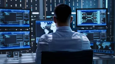 Person working at a desk with multiple monitors displaying data in a server room.