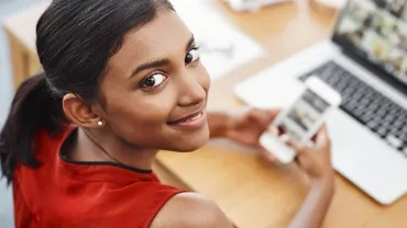 Woman in red top using smartphone and laptop at desk, smiling.