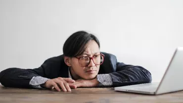 Person in glasses leaning on a table, looking at a laptop.