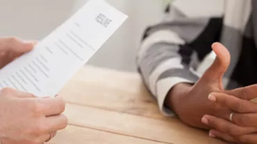 Close-up of an interview, one person holding a resume, another with folded hands on a wooden table.