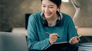 Woman with headphones studying at a desk with a laptop and notebook.