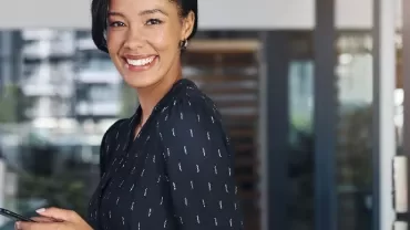 Woman in a black dress smiling and holding a phone in an office setting.