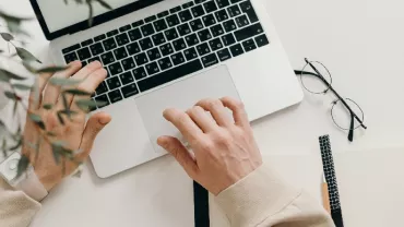 Person typing on a laptop with a notebook, pen, and glasses on a white desk.