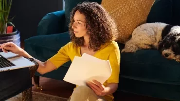 Woman working on a laptop with papers, sitting on the floor near a couch.