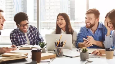 Group of young adults laughing and working together at a table with books, papers, and laptops.