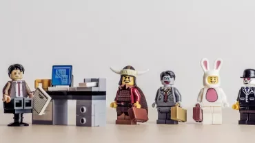LEGO figures in costumes stand in a line, one sits at a desk with a paper, depicting a quirky office scene.