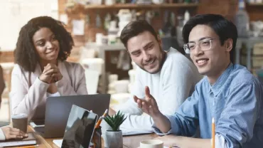 Diverse group of people in a casual meeting, discussing and smiling around a table with laptops.