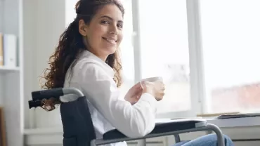 Woman in wheelchair smiling, holding a coffee cup next to a laptop.