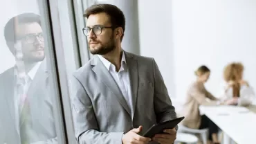 Businessman holding a tablet looks out the window; blurred meeting in the background.