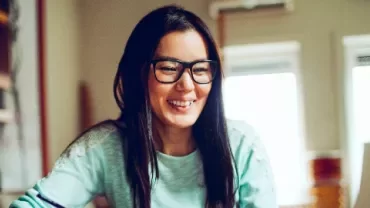 A woman with glasses smiling while working on a laptop at home.