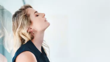 Woman leaning against a glass wall, eyes closed in thought.
