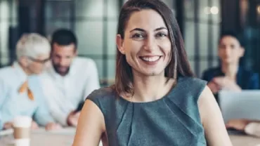 Smiling woman in a gray dress in an office setting with colleagues in the background.