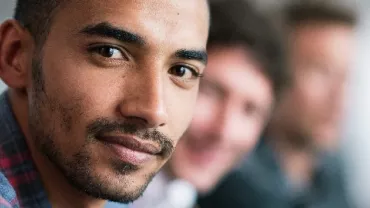 Smiling man in a plaid shirt, two people blurred in the background.