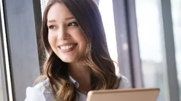 Woman smiling while holding a tablet by a window.