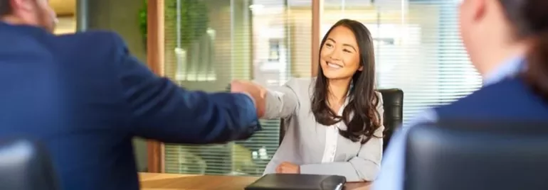 A woman smiles while shaking hands with a person across a table in an office setting.