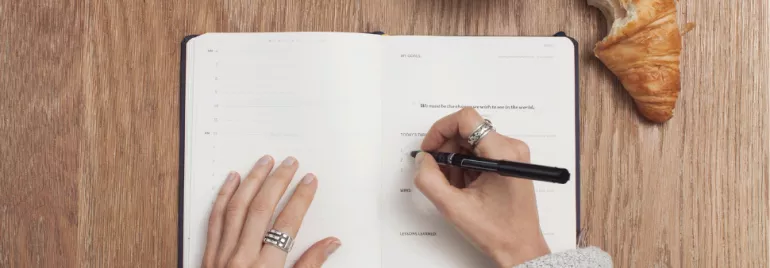 Person writing in a notebook on a wooden table with a croissant and coffee nearby.