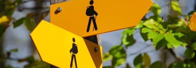 Two yellow hiking trail signs on a post with green leaves in the background.