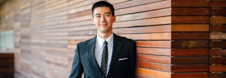 Man in a suit leaning against a wooden wall, looking at the camera.