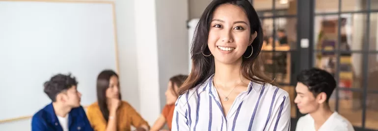 Smiling woman in a striped shirt stands in front of a group of people in a modern office environment.