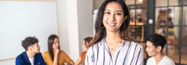 Smiling woman in a striped shirt stands in front of a group of people in a modern office environment.