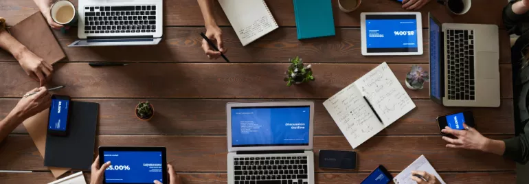 Top view of a group of people working with laptops, tablets, and notes around a wooden table.