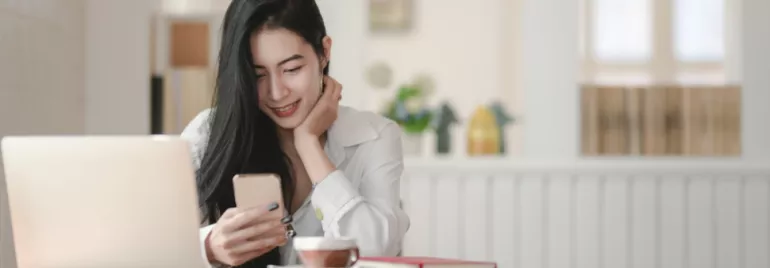 Woman smiling at phone in a cafe with a laptop and coffee.