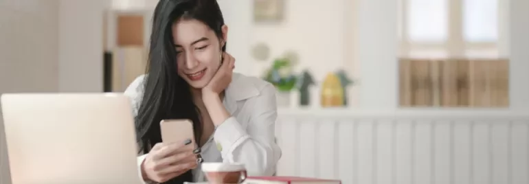 Woman smiling at phone in a cafe with a laptop and coffee.
