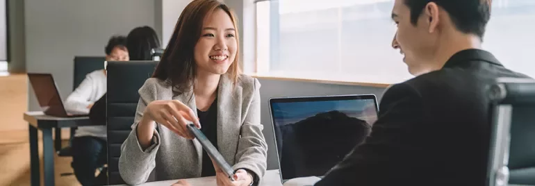 A woman is smiling and handing a document to a man in a modern office.