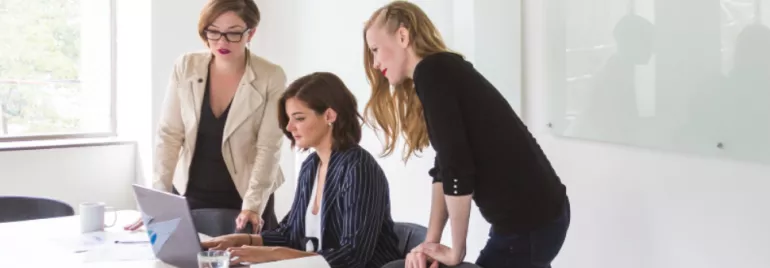 Three women discussing work around a laptop in a modern office.