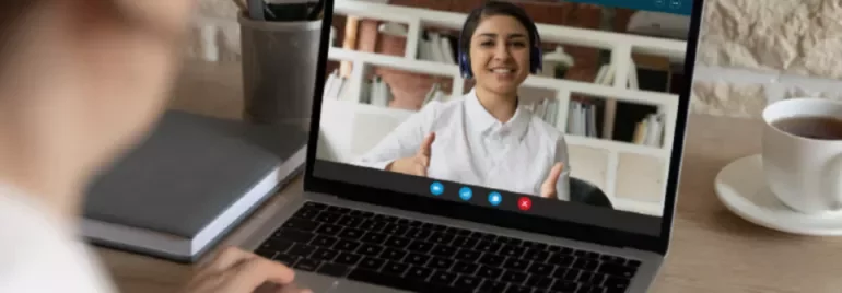 Person video calling on laptop at desk with coffee and notebooks.